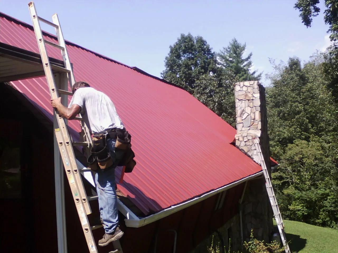 Roofer on a ladder beside a red metal roof and stone chimney — Soffit & Fascia Repair by Roofing Brothers in Fernwood